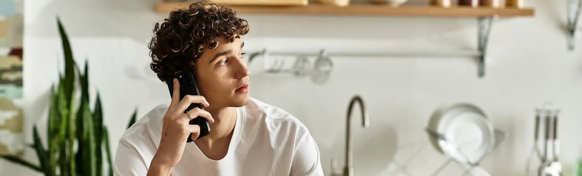 A young man with curly hair is talking on the phone in a modern kitchen while handling a car crash insurance call after a recent accident.