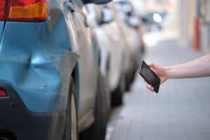 A person's hand holding a smartphone, photographing a damaged blue car bumper and taillight on a busy street, documenting hit-and-run collision evidence.