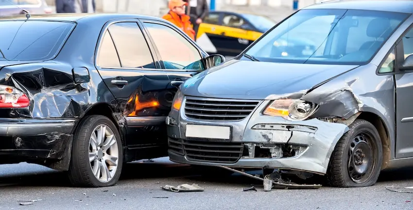 A black sedan with a severely caved-in rear passenger door and quarter panel is struck by a gray vehicle whose entire front bumper has been torn away and is hanging toward the pavement with plastic debris scattered across the wet urban road surface between the two damaged cars- a person in an orange jacket visible in the blurred background- and a yellow taxi partially visible behind them — the kind of violent urban collision commonly caused by distracted driving in Knoxville TN and could use legal help from Knoxville Car Accident Lawyer.