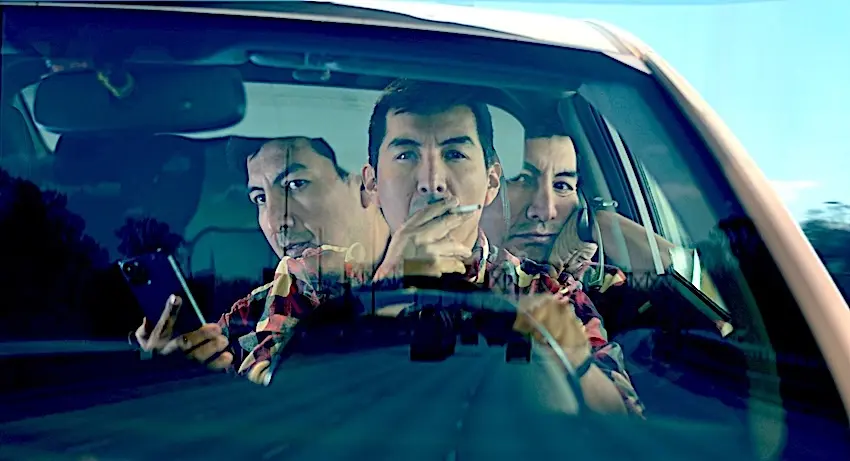 A composite multiple-exposure image shot through a vehicle's windshield shows the same male driver in a red plaid flannel shirt depicted three times simultaneously — once looking down at a dark smartphone in his left hand- once bringing a cigarette to his lips- and once staring forward at the road — against a bright blue sky and blurred roadside scenery- powerfully illustrating the dangerous range of behaviors that constitute distracted driving in Knoxville TN.