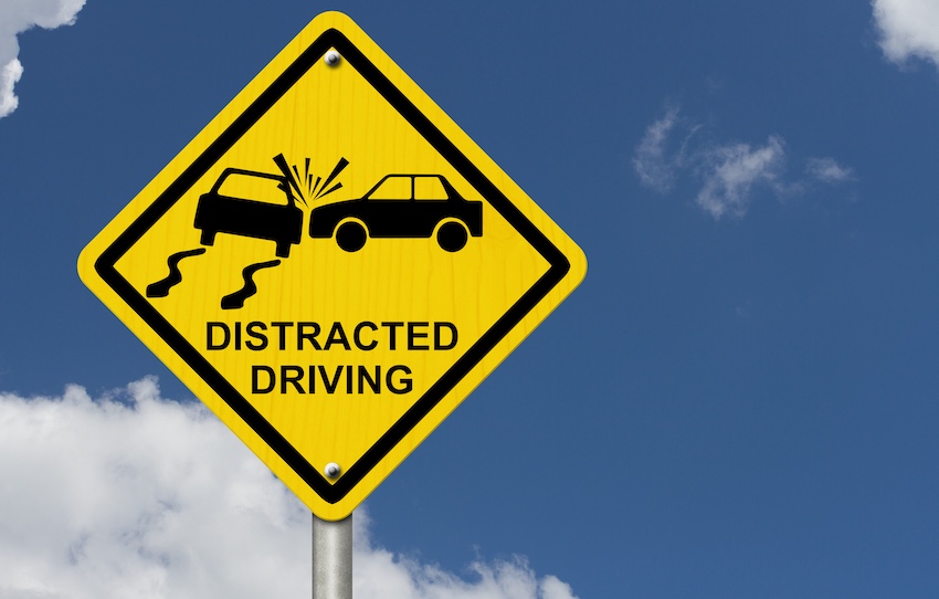 A bright yellow diamond-shaped warning sign mounted on a silver metal pole displays two black silhouetted vehicles in the middle of a rear-end collision with impact lines radiating from the point of contact- along with wavy skid mark lines beneath the striking car- and the bold black text "DISTRACTED DRIVING" printed across the lower half of the sign photographed against a vivid blue sky dotted with white clouds in the upper right corner — a roadside public safety warning about the dangers of distracted driving in Knoxville TN.